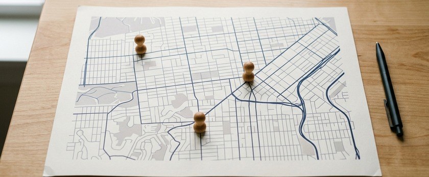 無地の市街地図の上に3本の木製ピンを立てた俯瞰イメージ。福岡入管の本局・西新分室・天神分室が別々の場所にあることを象徴。