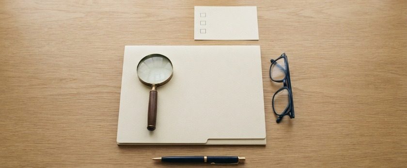 無地のフォルダの上に置かれたルーペと眼鏡、空欄のチェックカードの俯瞰イメージ。外部監査人による独立した点検を象徴。