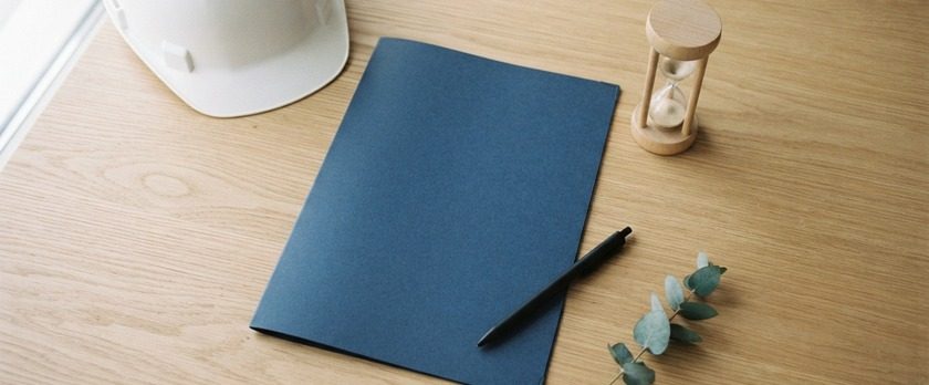 無地の書類フォルダと砂時計、白いヘルメットを並べた俯瞰イメージ。建設特定技能受入計画の認定に1か月半〜2か月かかることを象徴。