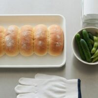 ステンレスの作業台に白いトレーのパンと野菜、白い手袋を並べた俯瞰イメージ。宮崎の特定技能・飲食料品製造業分野を象徴。