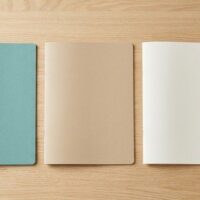机の上に無地のフォルダ3冊とペン、眼鏡、観葉植物を並べた俯瞰イメージ。事業所ごとに支援体制を整える実務を象徴。