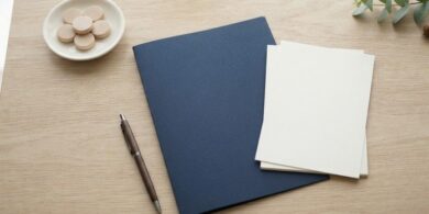 木の机に無地の紺色の書類フォルダと白紙の束、木製の丸い駒を並べた俯瞰イメージ。在留手数料の減額を求めるための疎明資料の準備を象徴。