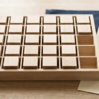 区画に分かれた無地の木製トレイに無地の木片が並び、右端の空いた区画のそばに予備の木片が置かれた俯瞰イメージ。分野の数は変わらず業務区分だけが増える案を象徴。