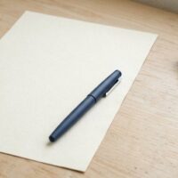 白紙の書類の上にペンが置かれ、少し離れたところに無地の木製印鑑が横向きに置かれた俯瞰イメージ。海外在住者は印鑑証明に代えて署名証明を使うことを象徴。
