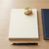 きちんと揃えた無地の書類の束に真鍮の文鎮を載せ、紺色のバインダーを添えた俯瞰イメージ。永住許可ガイドラインが確定したことを象徴。