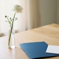 白い花を挿したガラスの花瓶と紺色のフォルダー、白い封筒を置いたイメージ。家族が亡くなった後の手続を象徴。
