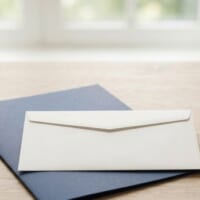 無地の白い封筒と紺色のフォルダー、小さな天びんを置いた机のイメージ。育成就労の給与と課税を象徴。