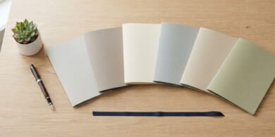 無地のフォルダを6つ並べ、紺色のリボンとペンを添えた机のイメージ。育成就労の協力覚書が6か国になったことを象徴。