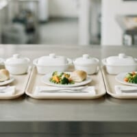 ステンレスの調理台に無地の食事トレーを3つ並べたイメージ。医療・福祉施設の給食を象徴。