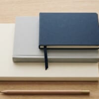 表紙が無地のノート3冊を階段のように少しずつずらして重ね、紺色のしおりひもを垂らした俯瞰イメージ。日本語能力の水準が一段上がることを象徴。