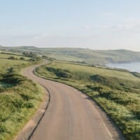朝の光の中、緑の丘と海沿いを通る車のいない道路のイメージ。地域の暮らしを支える運転の仕事を象徴。