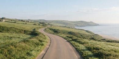 朝の光の中、緑の丘と海沿いを通る車のいない道路のイメージ。地域の暮らしを支える運転の仕事を象徴。