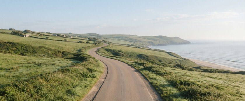 朝の光の中、緑の丘と海沿いを通る車のいない道路のイメージ。地域の暮らしを支える運転の仕事を象徴。